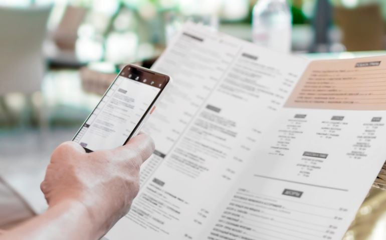 Person's hands holding menu and mobile phone in restaurant. A foreign man in cafe ordering food using word translation program while sitting at table. Mockup for smartphone screen