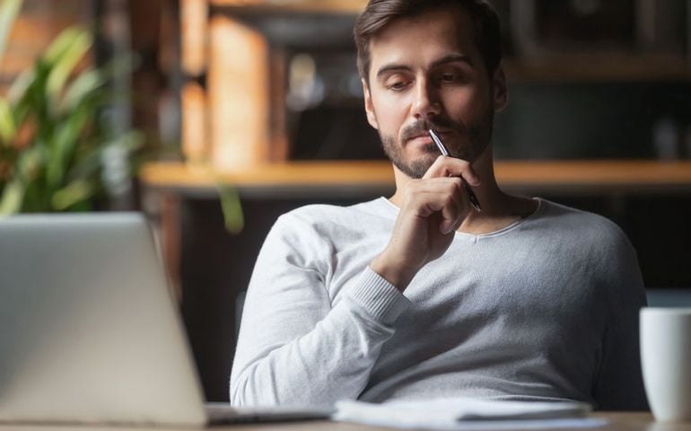 Pensive bearded man sitting at table drink coffee work at laptop thinking of problem solution, thoughtful male employee pondering considering idea looking at computer screen making decision