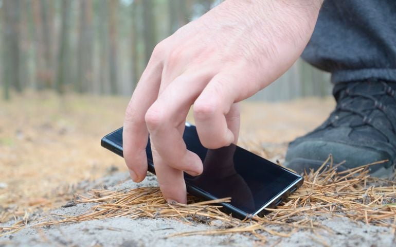 Male hand picking up lost mobile phone from a ground in autumn fir wood path. The concept of finding a valuable thing and good luck