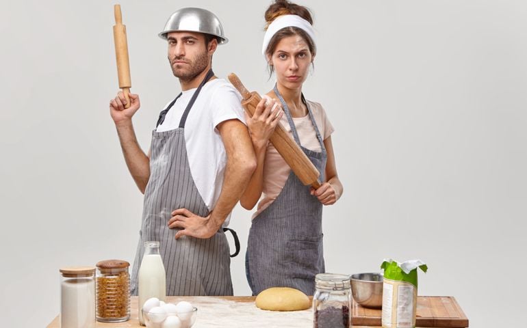 Serious woman and man stand back to each other, hold wooden rolling pins, participate in culinary battle, show who cooks better, compete in culinary arts, pose at kitchen. Cooking challenge.