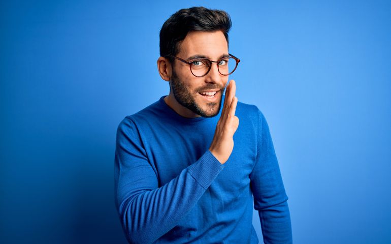 Young handsome man with beard wearing casual sweater and glasses over blue background hand on mouth telling secret rumor, whispering malicious talk conversation