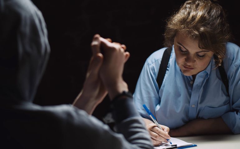 Female police officer interrogating criminal in handcuffs at desk in dark room. Woman cop questioning suspect and writing down information at police department