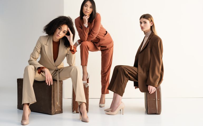 full length of stylish multicultural women in suits sitting on suitcases on white