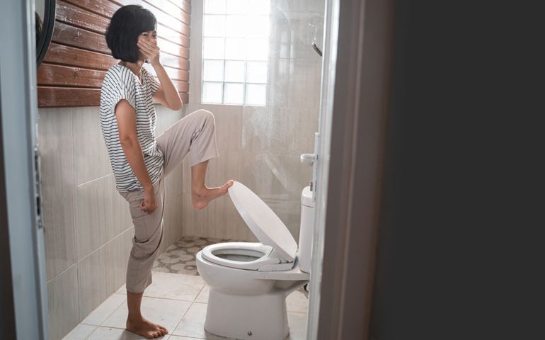 a woman cover her nose while opening the lid of the toilet with her feet in the shower