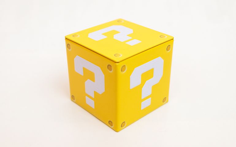 Indoor still-life photo of a yellow box with a big white question mark printed on each face. It recalls a graphic element of a famous platform video game.