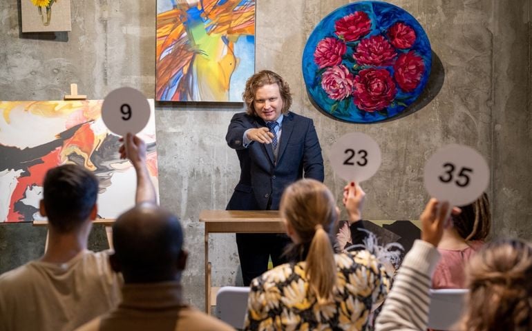 Mature male auctioneer in formalwear standing by tribune and pointing at one of people with auction paddles