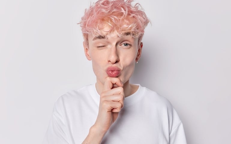 Handsome romantic man keeps lips folded hand on chin winks eye wants to kiss girfriend flirts with someone has pink wavy hair dressed in white t shirt poses indoor. Facial expressions concept