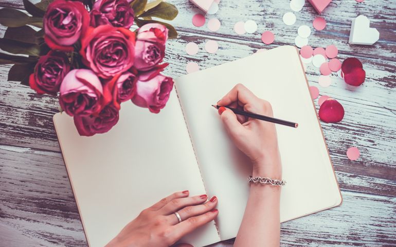 Female hands writing in open notebook and bouquet of roses on old wooden table. Top view. Toned image