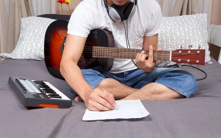 asian male composer, musician writing song & playing guitar on bed