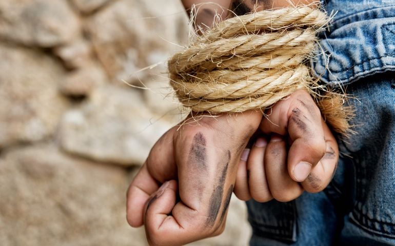 Dirty prisoner bound with rope. Hands behind back standing next to a wall.