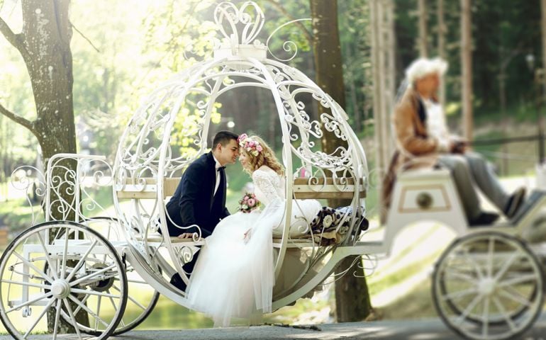 Handsome groom kissing blonde beautiful bride in magical fairy tale carriage in sunlit park