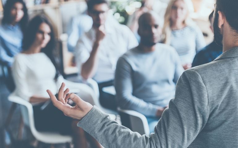 Business coach. Rear view of man gesturing with hand while standing against defocused group of people sitting at the chairs in front of him