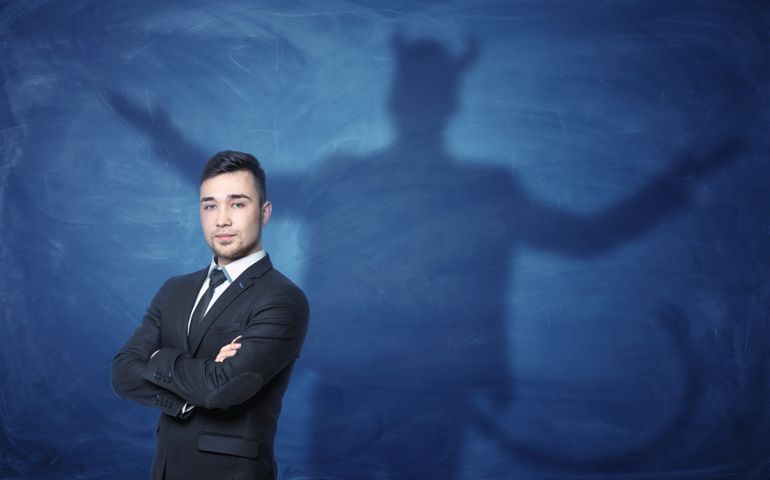 A businessman standing with his hands across and his shadow on a blue empty blackboard behind him with hands overspread, horns and a tail like a devil has. Self-confident behavior. Hidden feelings