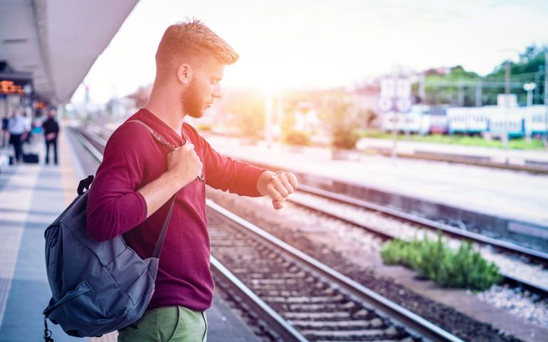 Young man checking the time on wristwatch at rail station platform - Student commuter waiting train at railway departures in pensive facial expression - Everyday lifestyle concept with sun halo filter