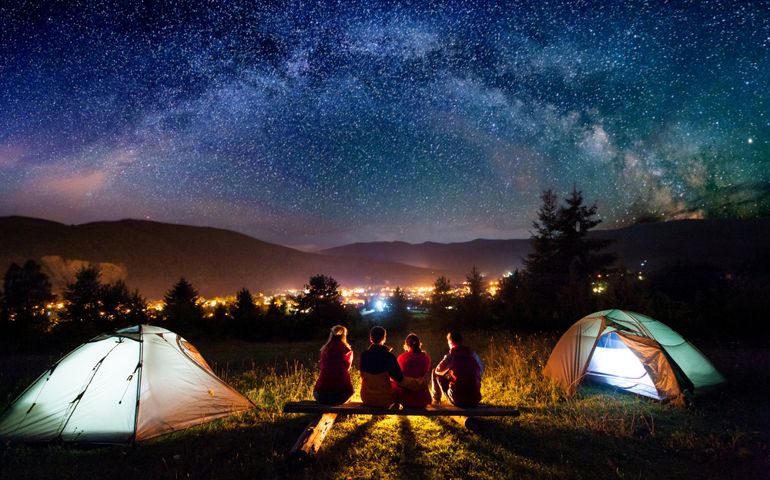 Friends hikers sitting on a bench made of logs and watching fire together beside camp and tents in the night. On the background beautiful starry sky, mountains and luminous town. Rear view