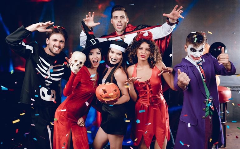 Halloween party. Six friends in costume cine villains posing with skull and pumpkin-lamp at a party. Around them flies confetti. They are having fun and laughing