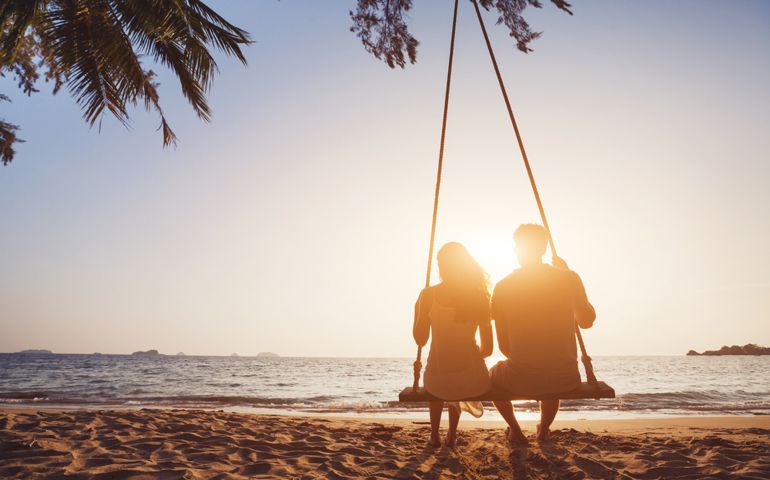 romantic couple in love sitting together on rope swing at sunset beach, silhouettes of young man and woman on holidays or honeymoon
