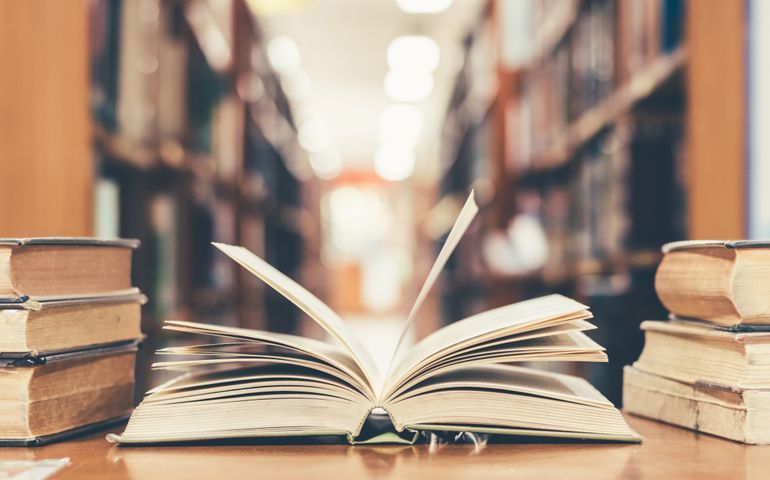 Education learning concept with opening book or textbook in old library, stack piles of literature text academic archive on reading desk and aisle of bookshelves in school study class room background