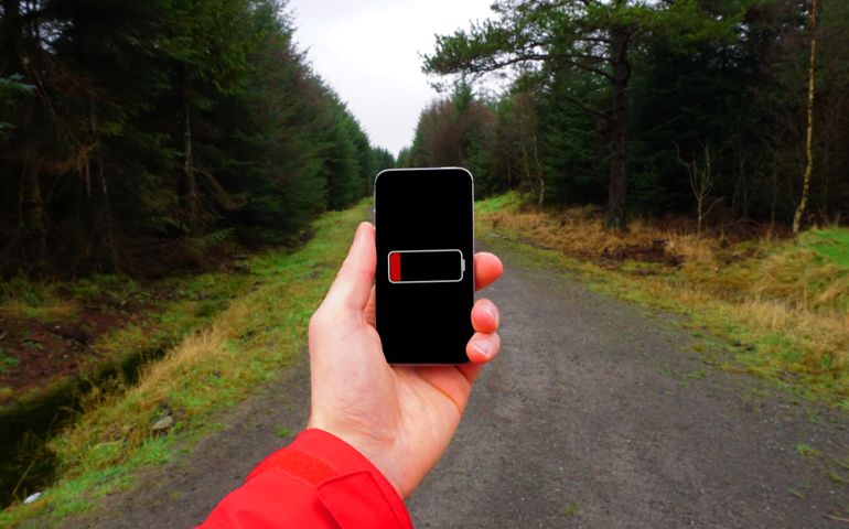 A person holding a phone in their hand with dead battery. It is cold and wet and in the background a trail runs through coniferous forest.