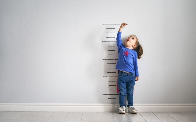 Little child is playing superhero. Kid is measuring the growth on the background of wall. Girl power concept.