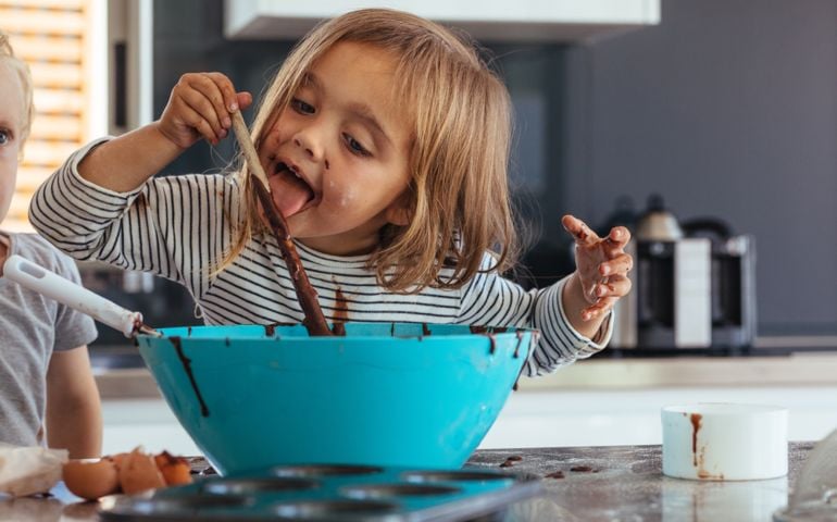 Little girl licking spoon while mixing batter for baking in kitchen and her brother standing by. Cute little children making batter for baking.