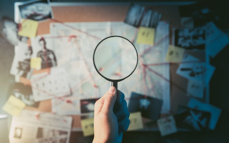 Detective hand holding a magnifying glass in front of a board with evidence, crime scene photos and map. high contrast image