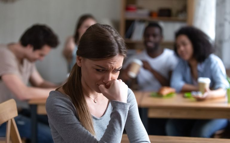 Diverse multi-ethnic friends sitting together in cafe talking having fun, focus on frustrated shy girl sitting separately by others teenagers feels unhappy because peers not accept her she is outcast