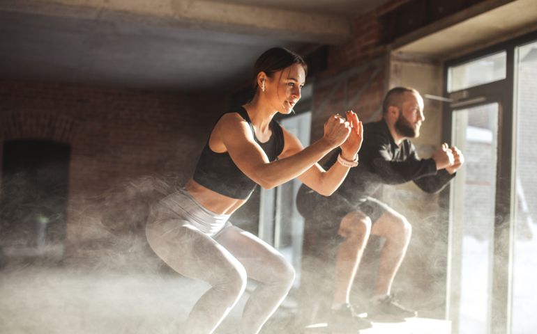 Fit caucasian couple doing squats on boxes in a crossfit style gym, well lit by bright sunlight.