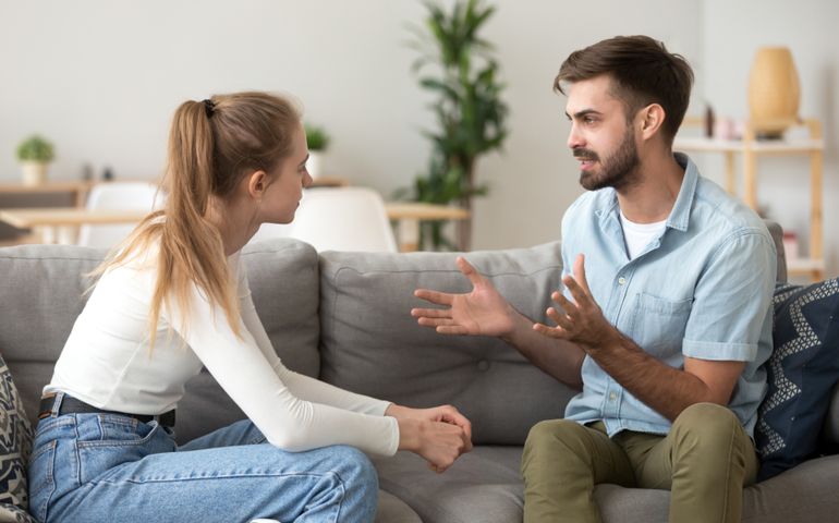 Serious young couple sitting together on sofa, talking about relationships, spending time together at home, focused wife listening to speaking husband, friends having conversation
