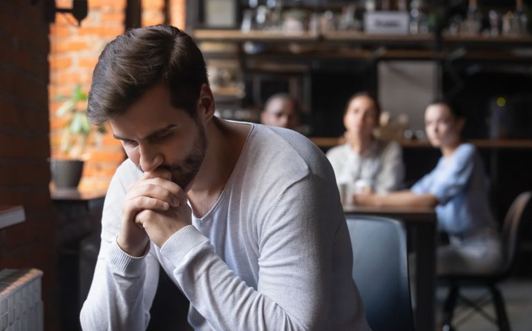 On foreground sad caucasian millennial guy outcast sitting separately from other diverse friends in cafe feels unhappy having communications difficulties, diffidence, jealousy, discrimination concept