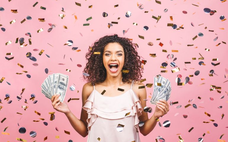 Black afro american girl won money. Happy young woman holding dollar currency satisfied isolated over pink background with confetti.