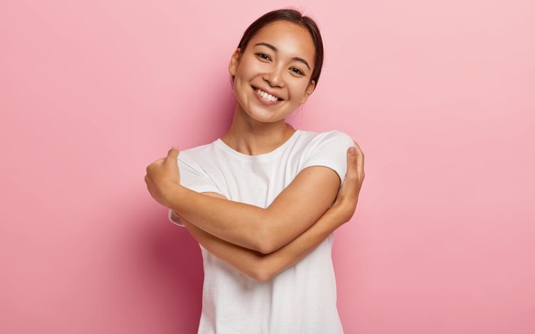Love yourself. Pretty glad Asian girl embraces herself, feels comfort and care, tilts head, wears white t shirt, has no make up, isolated over rosy wall, thinks about lover, wants be in his warm arms