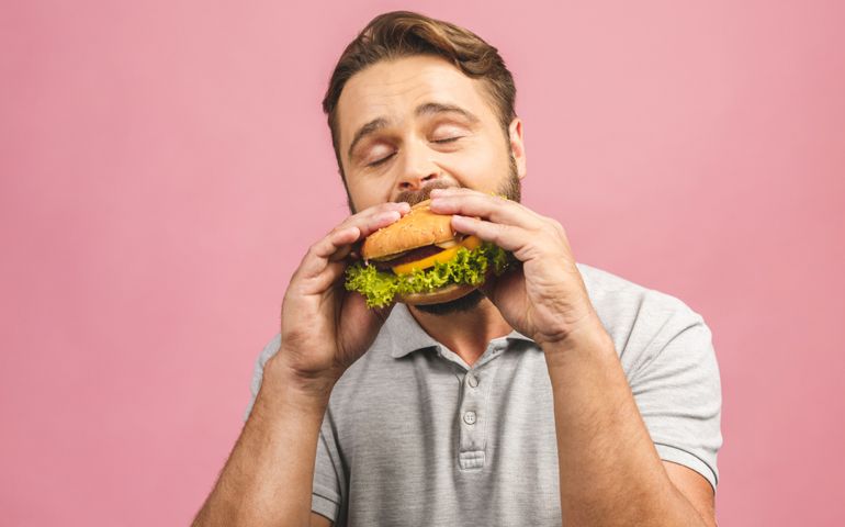 Young man holding a piece of hamburger. Bearded gyu eats fast food. Burger is not helpful food. Very hungry guy. Diet concept. Isolated over pink background.