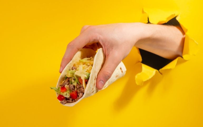 Hand holding a taco through a torn hole in yellow paper background. Hot price, discount on favorite Mexican dishes.