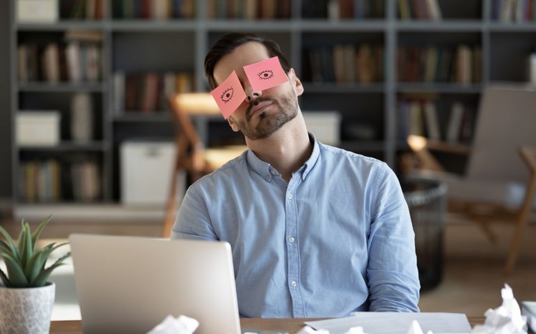 Exhausted tired businessman with painted eyes on stickers, adhesive notes on face sleeping at workplace, sitting at desk with laptop, unproductive lazy young male dozing, working on difficult project