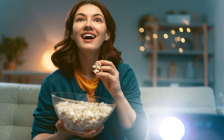 Young woman watching projector, TV, movies with popcorn in the evening. Girl spending time at home.
