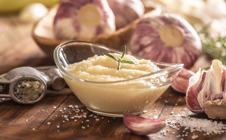 Garlic paste in a glass bowl with peeled garlic, salt and garlic heads.
