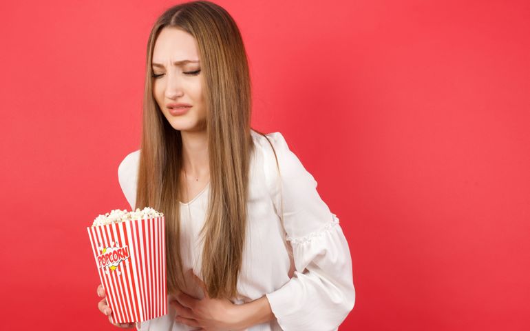 Young caucasian woman holding bucket with popcorn standing over isolated red background with hand on stomach because nausea, painful disease feeling unwell. Ache concept.