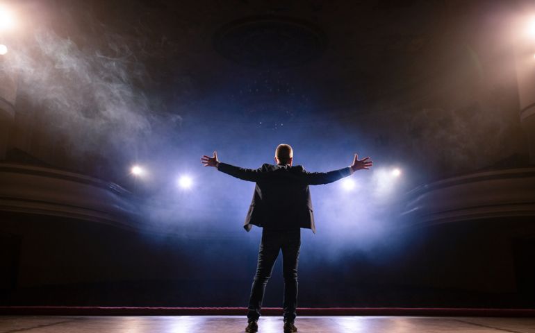 Showman. Middle Age Male entertainer, presenter or actor on stage. Arms to sides, smoke on background of spotlight. Rear view portrait of a male public speaker speaking at the microphone, pointing