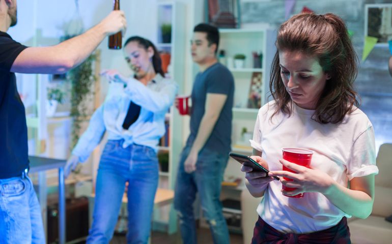 Shy young woman using her smartphone while her friends are dancing.