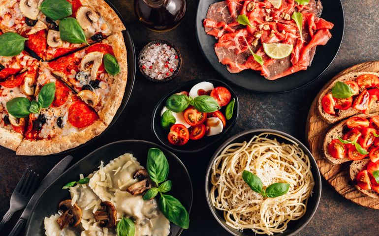 Full table of italian meals on plates Pizza, pasta, ravioli, carpaccio. caprese salad and tomato bruschetta on black background. Top view