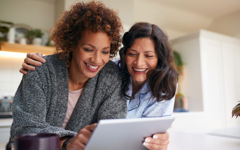 Same Sex Mature Female Couple At Home In Kitchen In Pyjamas Drinking Coffee With Digital Tablet