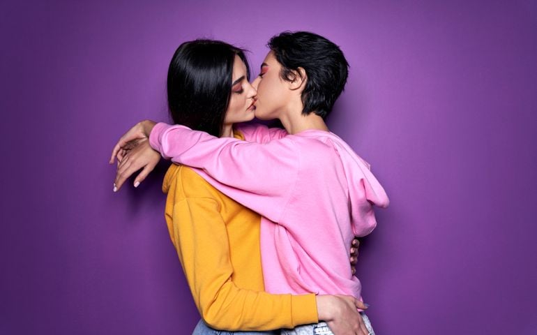 Two pretty sexy stylish cool generation z girls lgbtq lesbian couple dating in love hugging enjoying intimate tender sensual moment together kissing with eyes closed isolated on purple background.