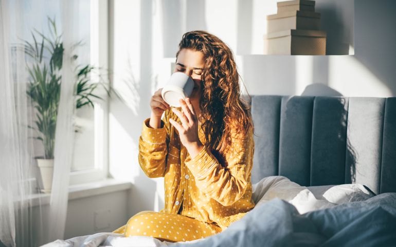 Woman in yellow pajama sitting on bed and drinking coffee at the sunny morning bedroom.