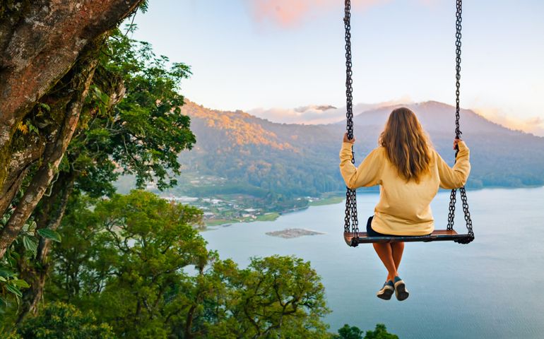 Summer vacation. Young woman sit on tree rope swing on high cliff above tropical lake. Happy girl looking at amazing jungle view. Buyan lake is popular travel destinations in Bali island, Indonesia