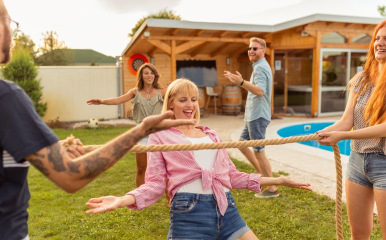 Group of cheerful young friends having fun at summertime outdoor party by the swimming pool, participating in limbo dance contest, passing below the rope while dancing