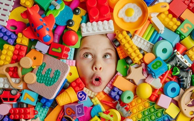 Funny surprised little girl lying in the chaos of toys. Kid's face surrounded by building blocks.