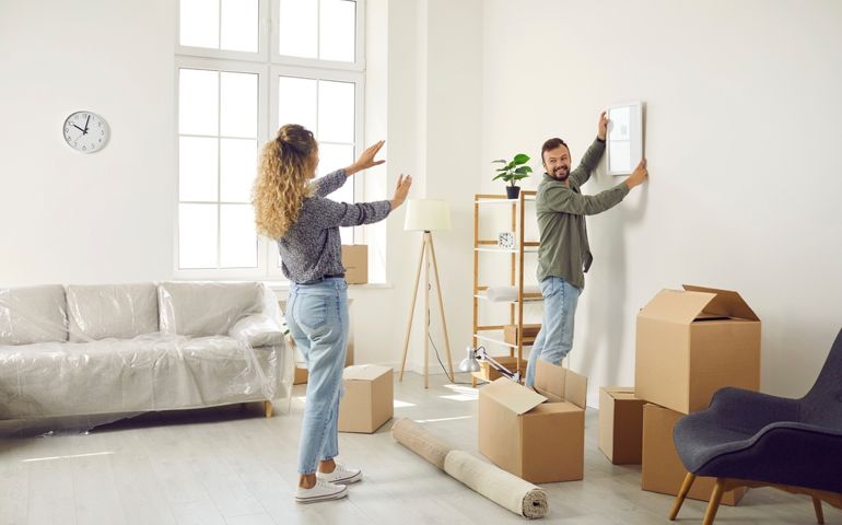 Happy married couple who recently bought house are unpacking stuff and decorating new home. Young man and woman are making living room cozy and choosing place on wall to hang painting or family photo