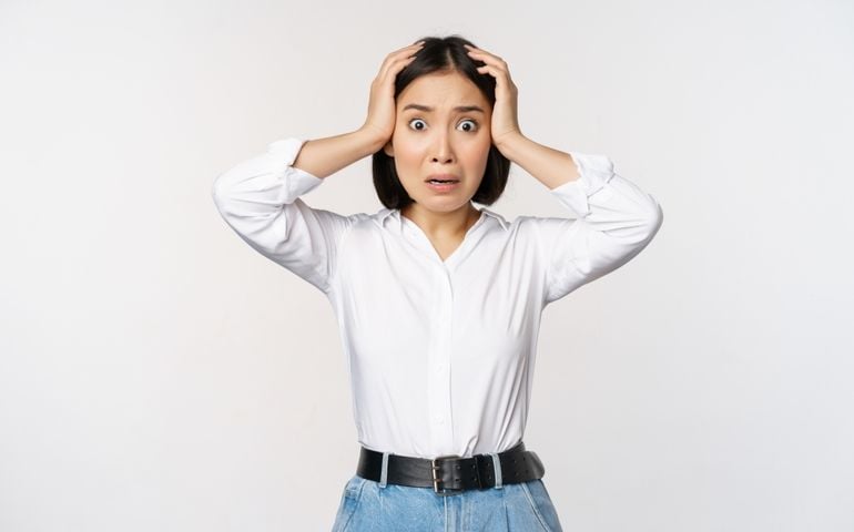 Image of shocked anxious asian woman in panic, holding hands on head and worrying, standing frustrated and scared against white background
