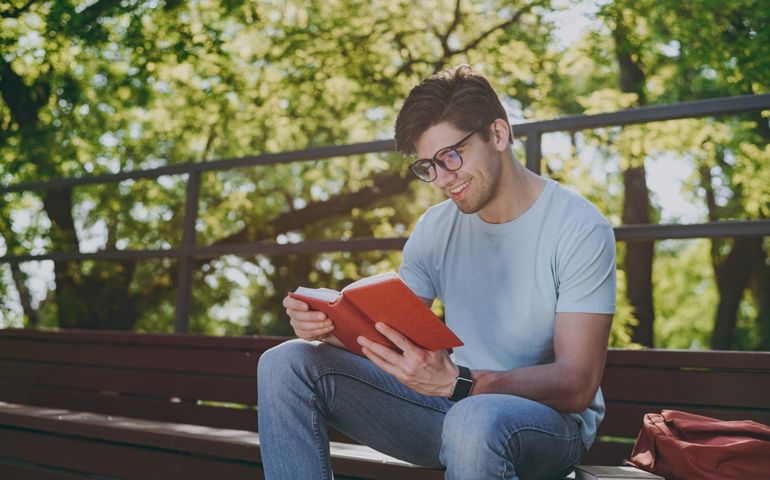 Young sunlit student man 20s wearing blue t-shirt eyeglasses backpack read book sitting on bench walking rest relax in sunshine spring green city park outdoors on nature. Education high school concept